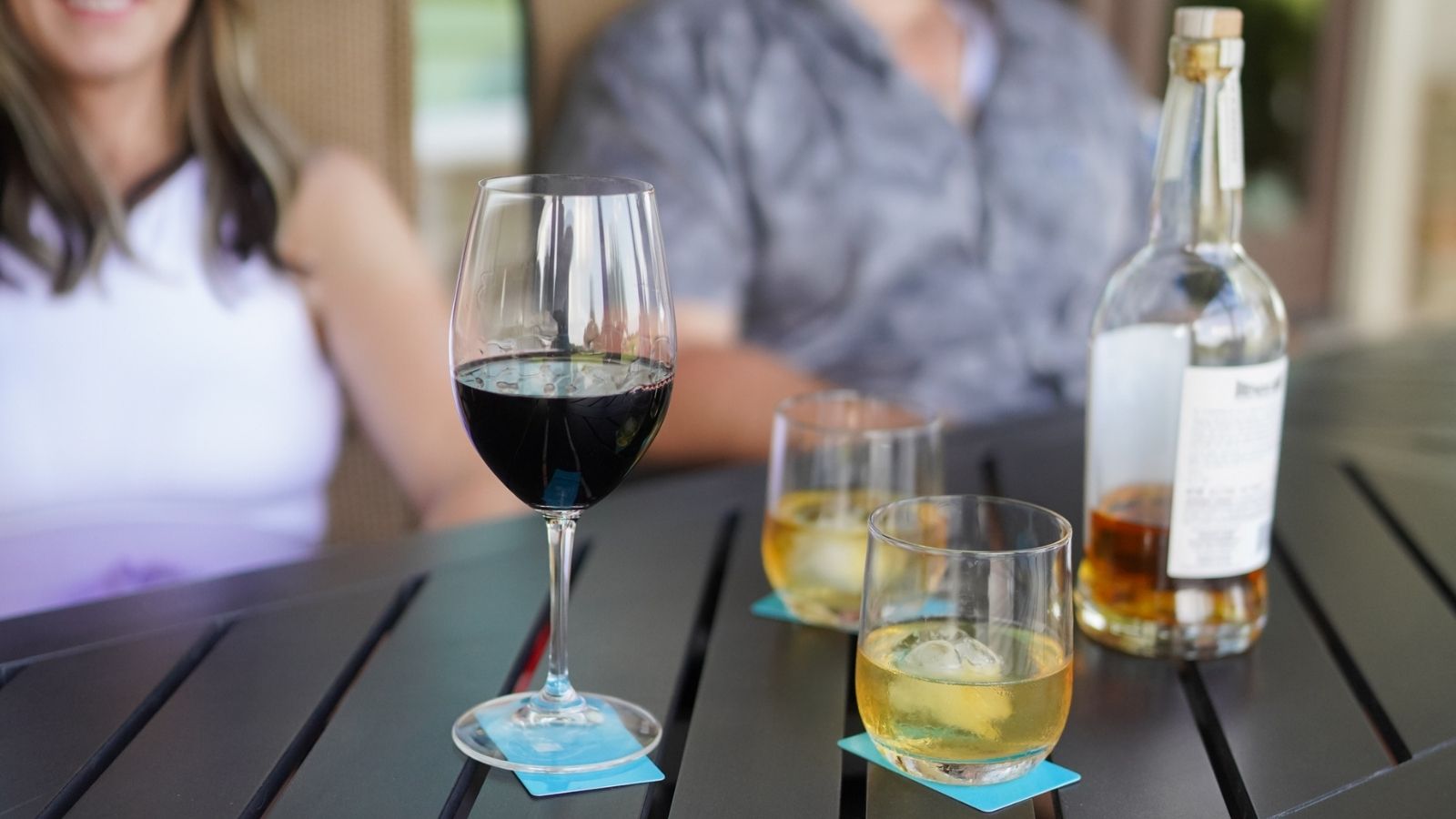 Two people sitting at a table with wine glasses, a bottle of wine, and bottle of whiskey. Glasses are sitting on BOCE coaster cards