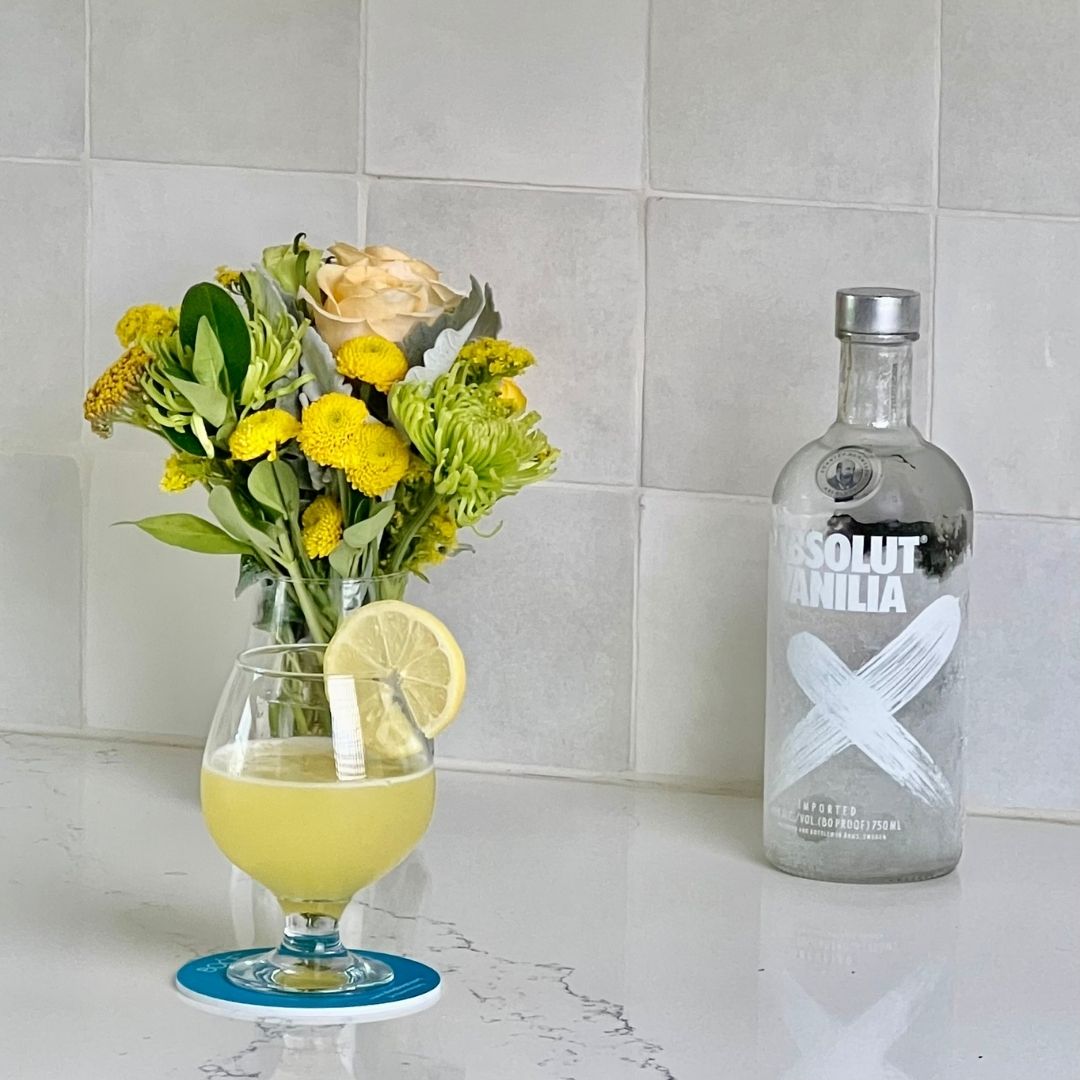 Bottle of Absolut Vanilia vodka with a glass of lemonade and flowers on a tiled surface with the glass resting on a BOCE coaster