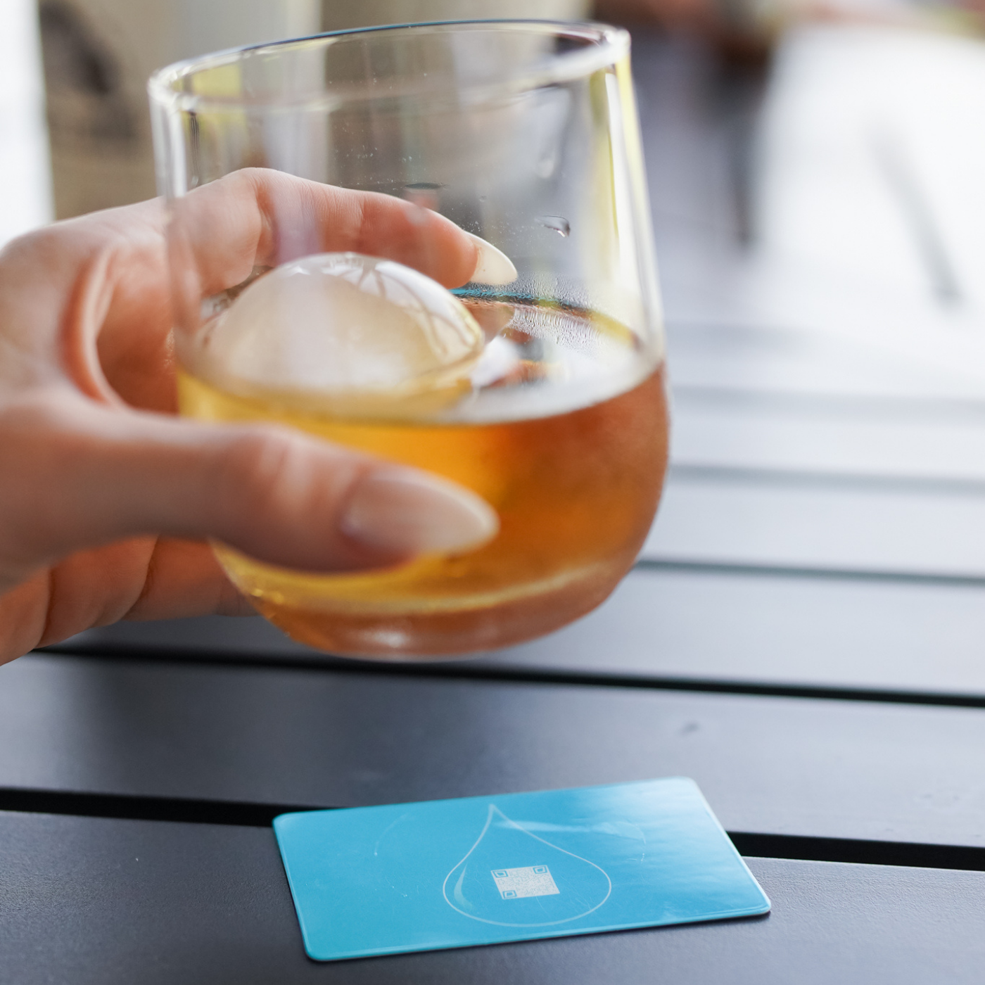 Hand holding a glass of amber liquid with ice, placed on a wooden surface next to a BOCE blue card.