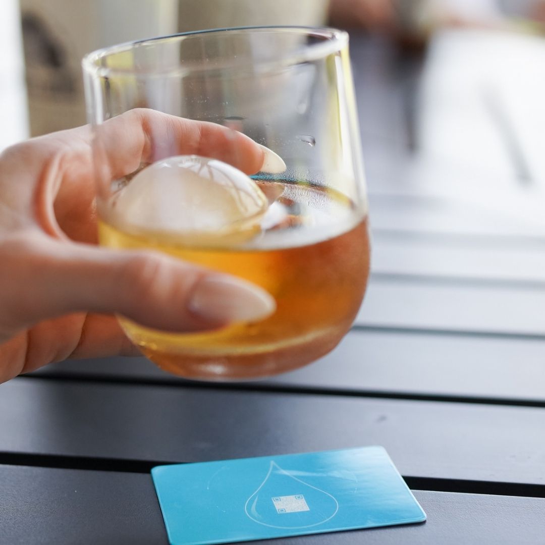 Hand holding a glass of whiskey with a blurred background placing the drink on a BOCE card coaster
